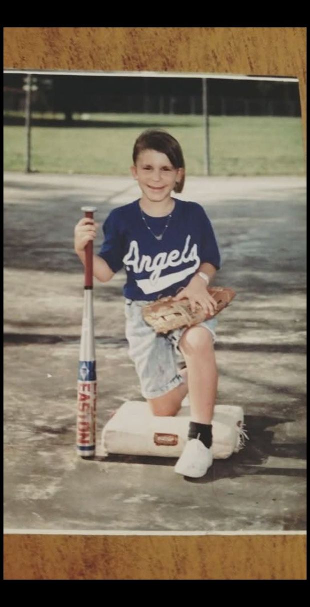 An old print of a child in a blue Angels shirt posed with a bat beside a base, photographed against wood paneling