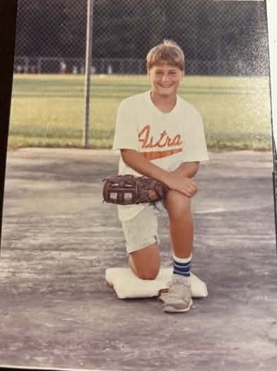 A young player kneeling on the pavement in a white Astros shirt and tube socks, a leather glove resting on one knee