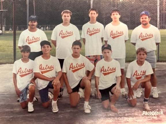 A faded team photograph of players in white Astros shirts, one row kneeling and one row standing with two adults, in front of a chain link fence