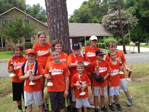 A team of children in orange Astros shirts holding trophies beside a pine tree, with two adults standing among them