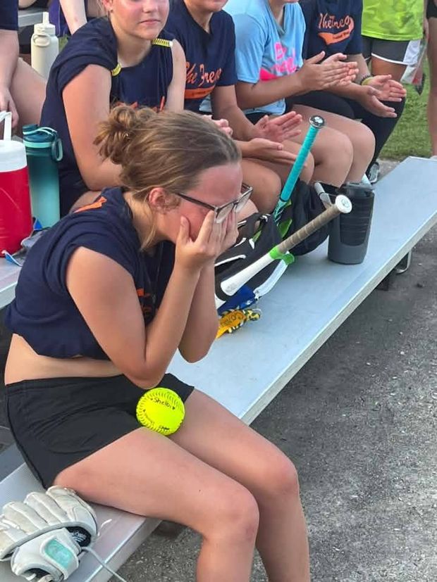 A girl sitting on the bench with a softball beside her while teammates clap behind her