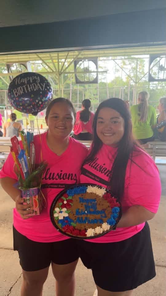 Two players in Illusions shirts holding a decorated cookie cake and a birthday balloon under the pavilion