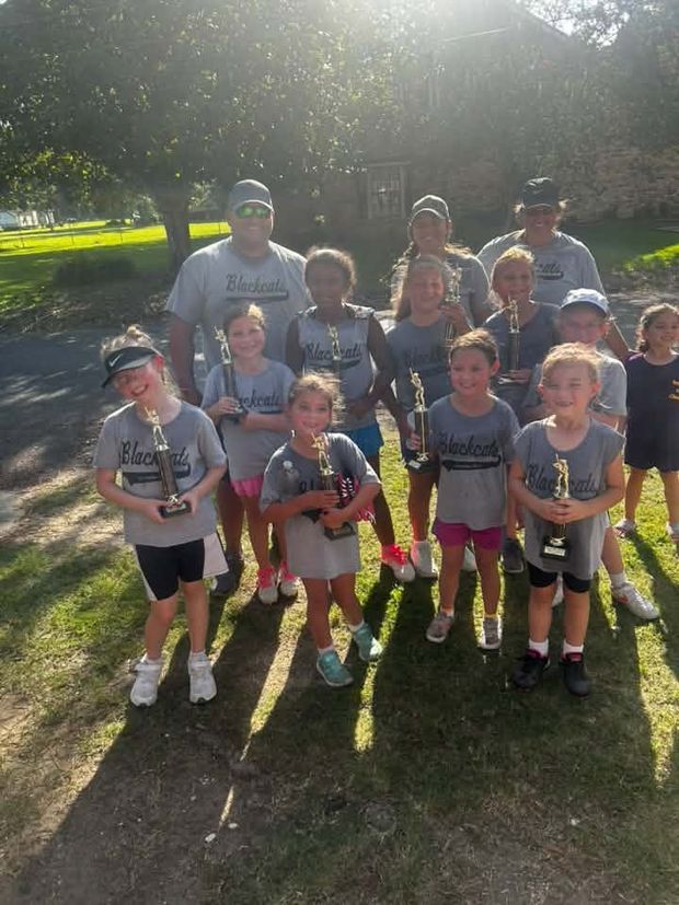 A team in grey Blackcats shirts standing on the grass in low evening light holding trophies