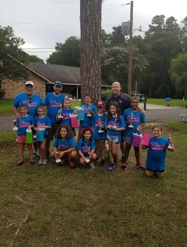 A girls team in blue shirts with their coaches holding cups beside a pine tree