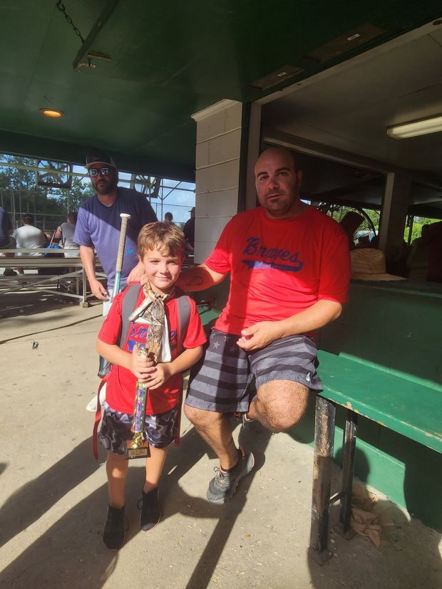 A man kneeling beside a boy in matching red Braves shirts, the boy holding a trophy