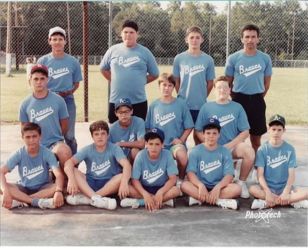 A posed team photograph of players and coaches in light blue Braves shirts, arranged in three rows in front of the outfield fence