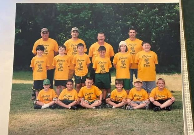 A printed team photograph of players in gold Cajun Boys shirts standing and kneeling on grass in front of trees