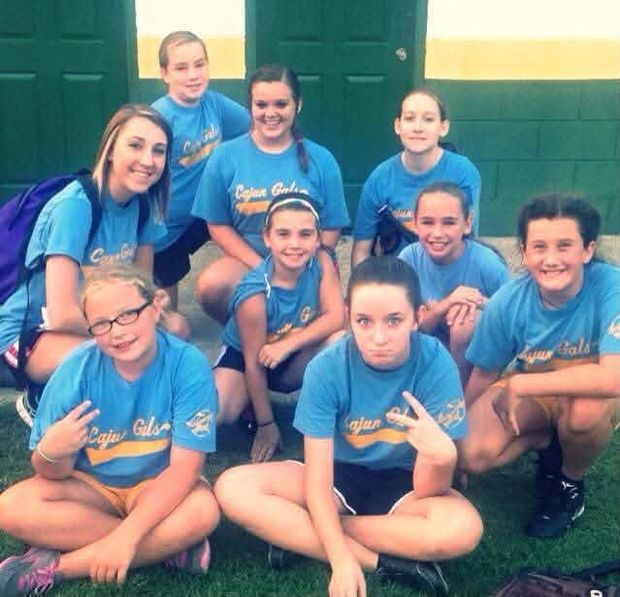 A girls team in blue Cajun Gals shirts posed in front of the green dugout doors