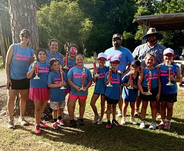 A team in blue Cajun Girls shirts with adult coaches, holding red trophies under pine trees