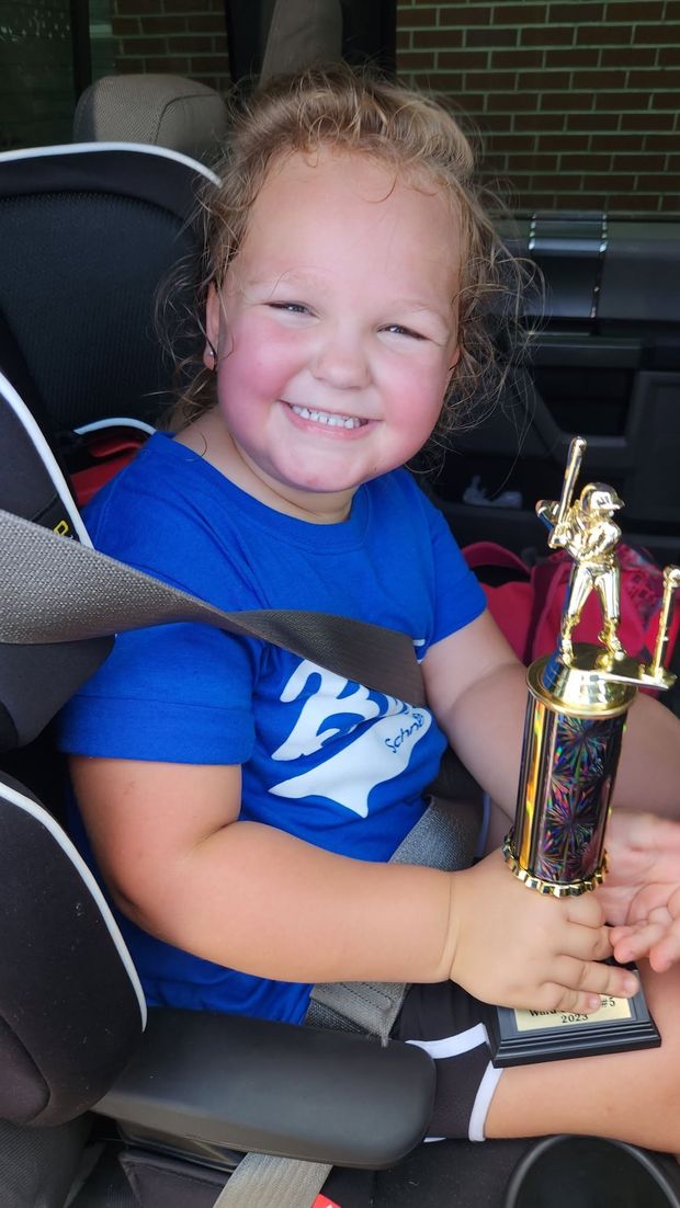 A smiling toddler in a blue shirt buckled into a car seat holding a trophy