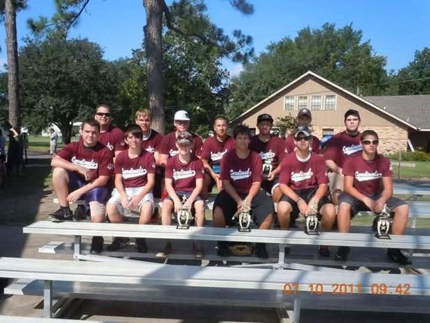 A team in maroon Cardinals shirts sitting on bleachers with trophies, with a camera datestamp in the corner
