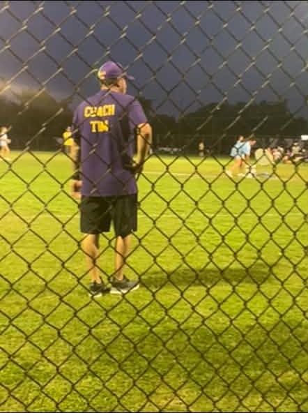 A coach in a purple shirt standing behind the outfield fence under the lights
