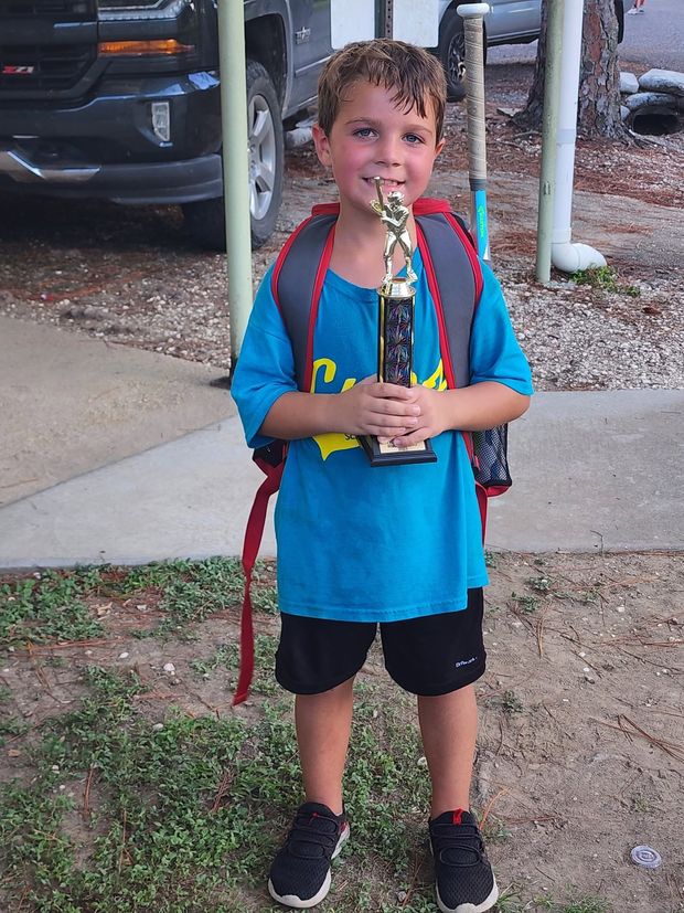 A boy in a blue Cubs shirt wearing a backpack and holding a trophy