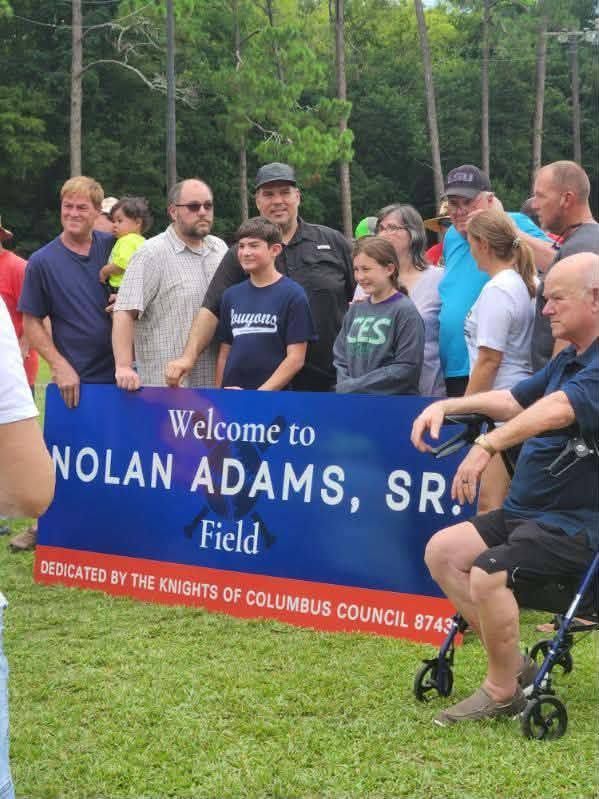 A crowd gathered on the field around a blue banner reading Welcome to Nolan Adams Sr Field