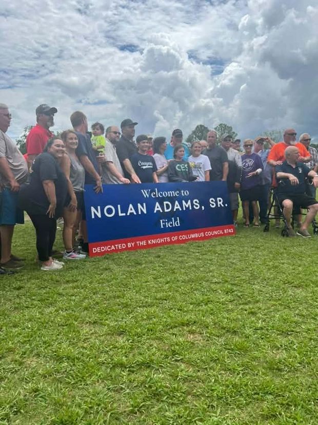 A large group posed behind the Welcome to Nolan Adams Sr Field banner under a cloudy sky