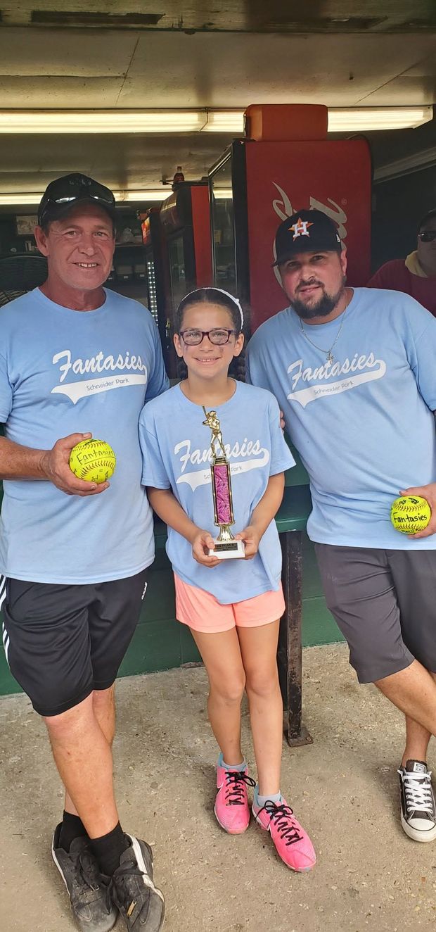 A girl in a light blue Fantasies shirt holding a trophy between two coaches holding softballs