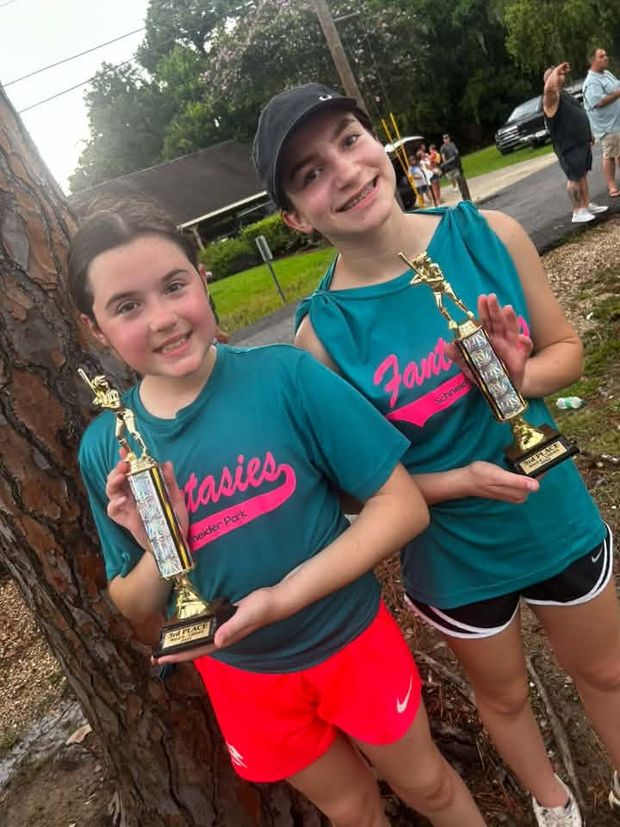 Two girls in teal Fantasies shirts holding trophies beside a pine tree