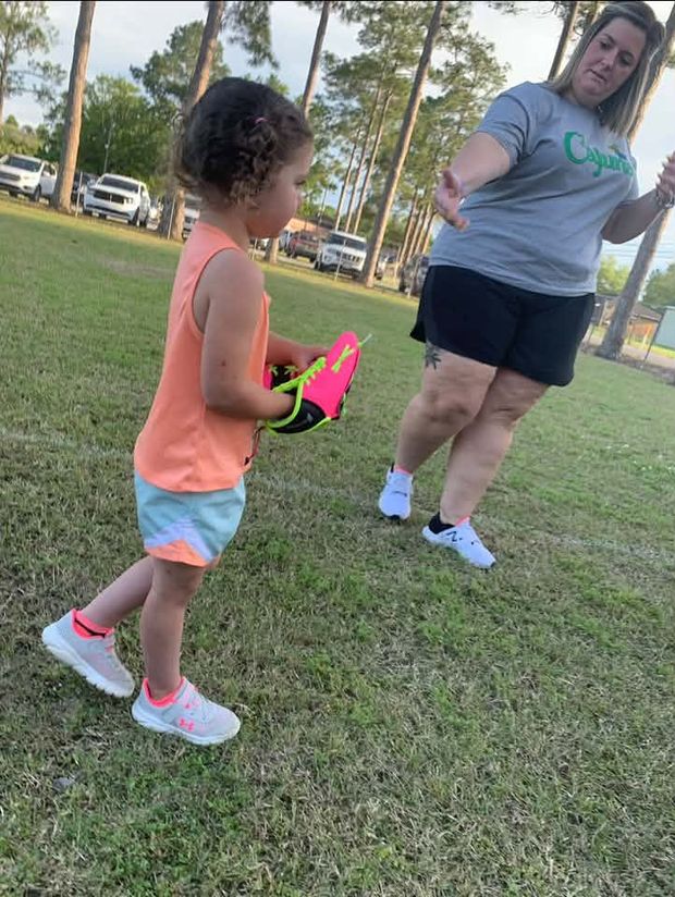 A toddler holding a glove while a woman in a Cajuns shirt reaches toward her on the grass
