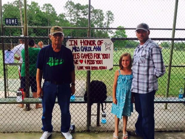 A hand-made sign honoring Mrs Carol Ann hung at the backstop, with a girl and two men standing beside it