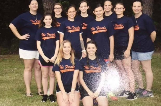 A softball team in navy Freaks shirts standing and kneeling together on the grass in front of trees