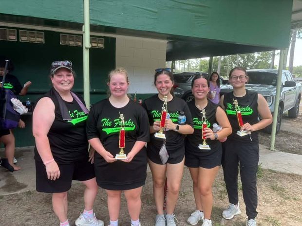 Four players in black Freaks shirts holding trophies beside an adult, standing under the covered dugout