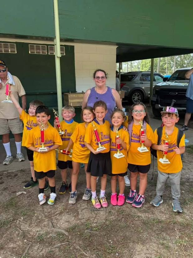 Small children in gold shirts holding red trophies near the dugout