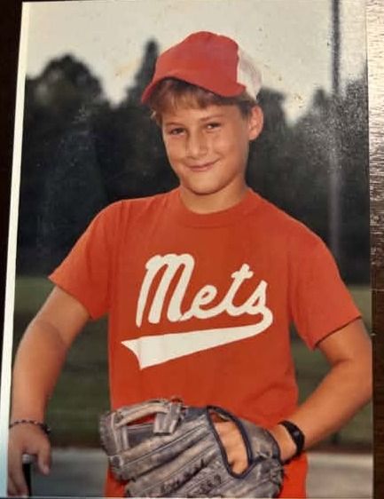 A young player in a red Mets shirt and red cap holding a well worn leather glove