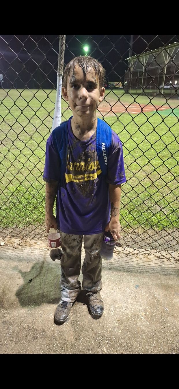 A young player soaked and covered in mud standing by the fence at night