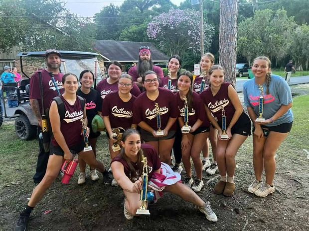 A girls team in maroon Outlaws shirts holding tall trophies with a golf cart behind them