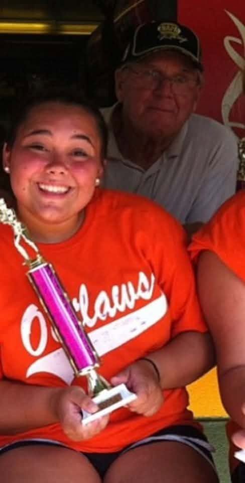 A girl in an orange Outlaws shirt holding a pink trophy beside an older man in a cap