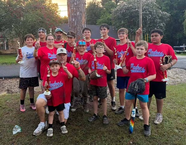 A team in red Padres shirts holding trophies under a pine tree