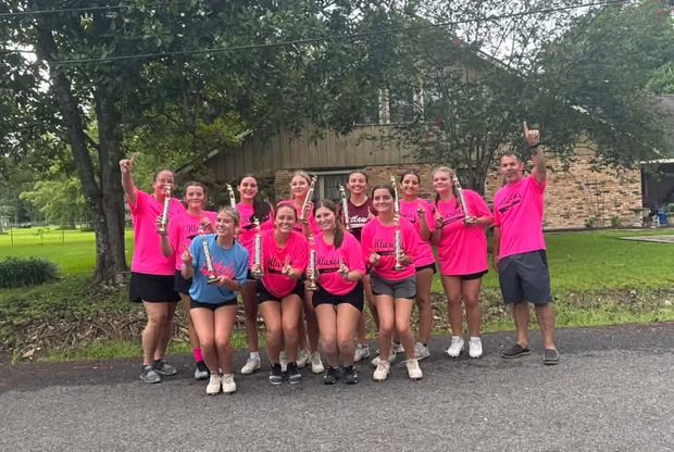 A softball team in pink shirts holding trophies on the grass, with their coach standing at one end