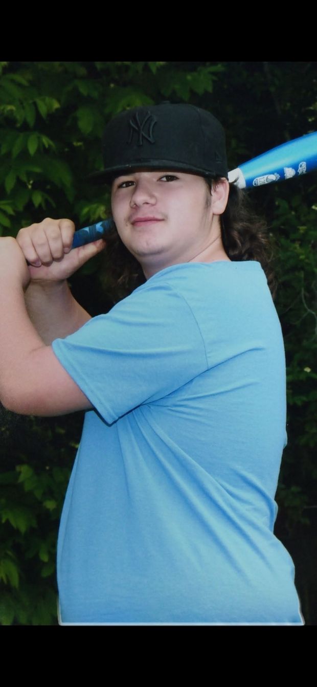 A teenager in a light blue shirt and dark cap in a batting pose