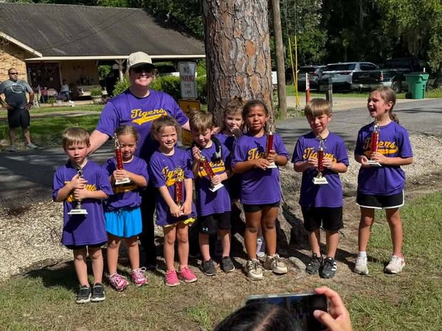 A team of young children in purple shirts holding trophies under a pine tree, with a coach standing behind them