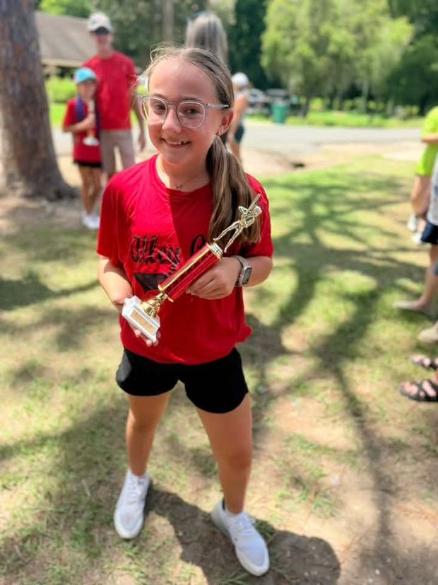 A girl in a red shirt holding a trophy under the trees