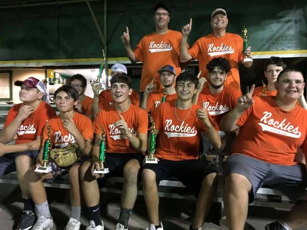 Teenage players in orange Rockies shirts sitting on bleachers at night with trophies