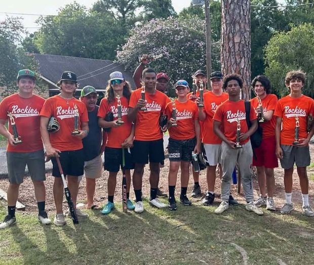 Teenage players in red and orange Rockies shirts holding trophies near the trees