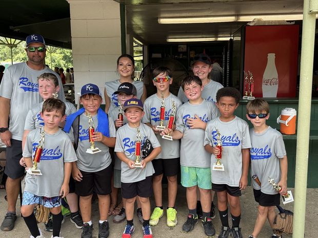 A team in grey Royals shirts holding red trophies in front of the concession stand