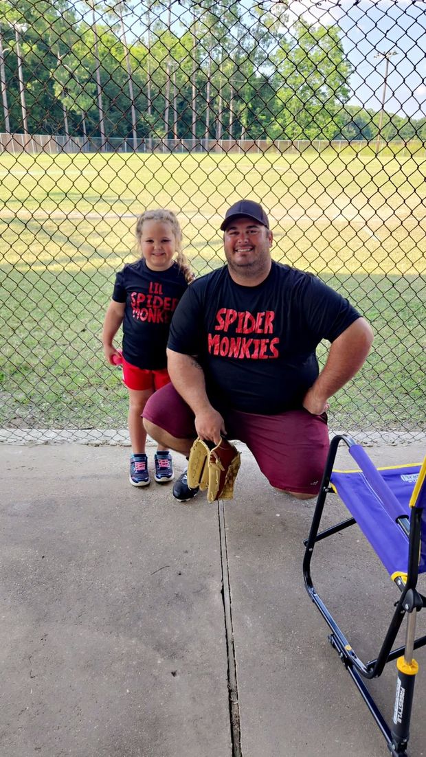 A man kneeling beside a young girl in matching Spider Monkies shirts by the fence