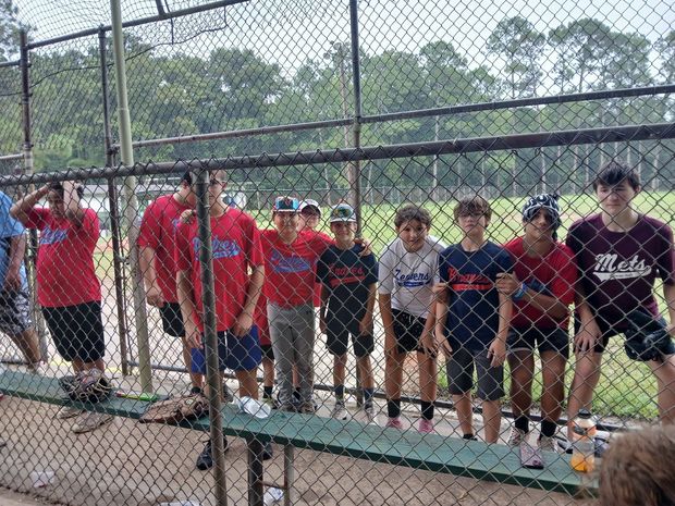 A row of players in red and navy baseball jerseys standing along a chain link fence beside the field