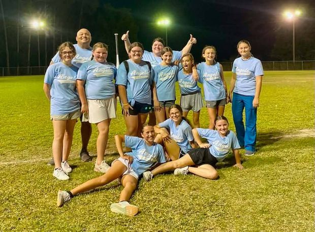 A team in light blue shirts standing and sitting on the outfield grass at night with the field lights on behind them