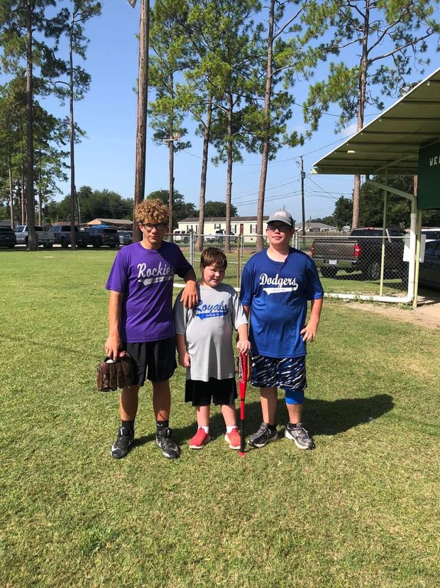 Three young players standing on the grass with bats and a glove, two in blue shirts and one in purple