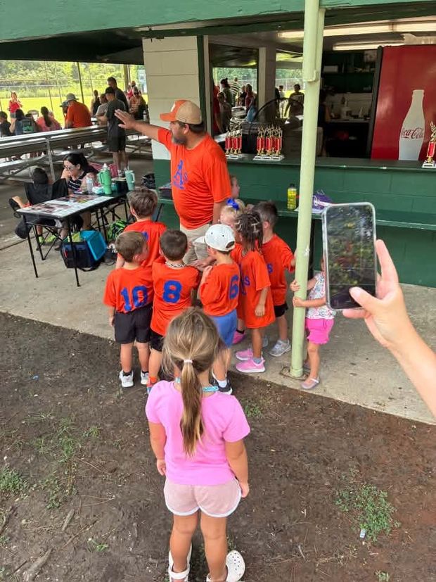 Small children in orange shirts gathered at the trophy table while a coach directs them