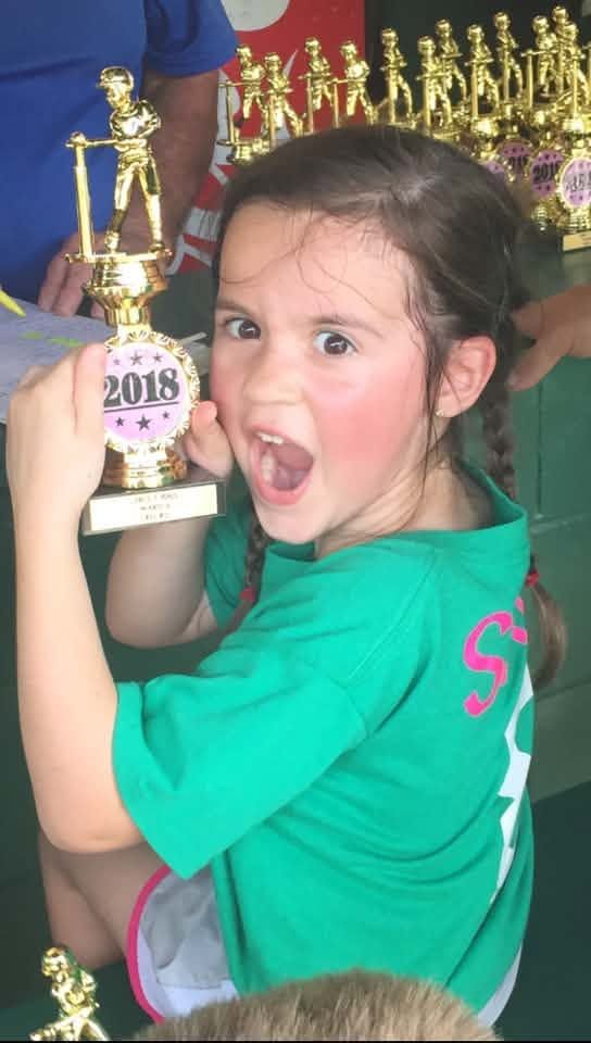 A girl in a green team shirt holding a trophy marked 2018, with the trophy table behind her