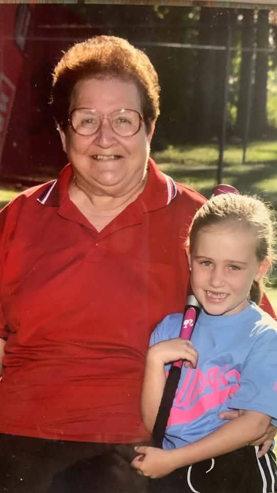 An older woman in a red shirt with her arm around a young player in a light blue team shirt who is holding a bat