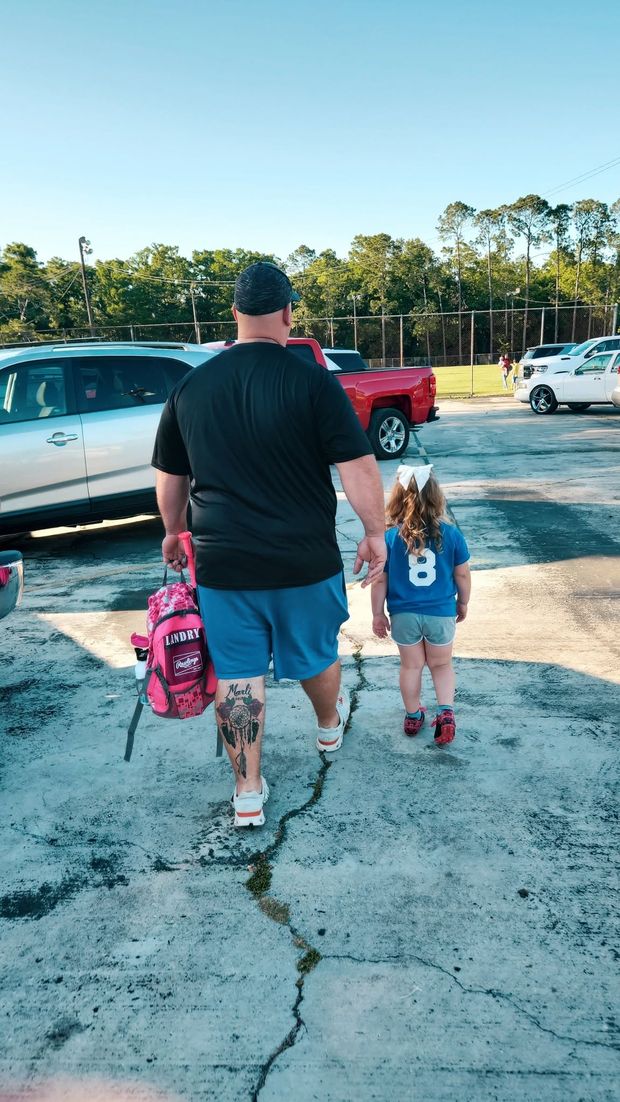 A man and a small child in a numbered jersey with a backpack walking toward the field