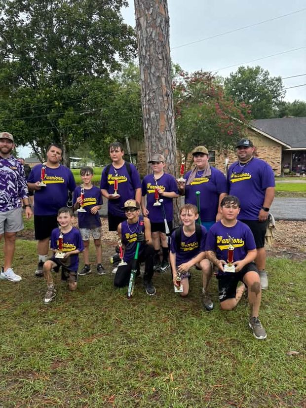 Children in purple Warriors shirts holding trophies with adult coaches beside a pine tree