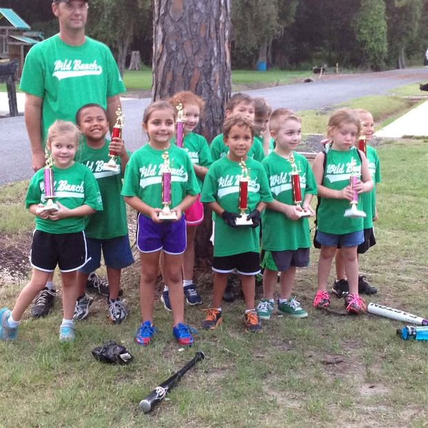 A team of small children in green Wild Bunch shirts holding trophies under a pine tree, with a coach standing behind them