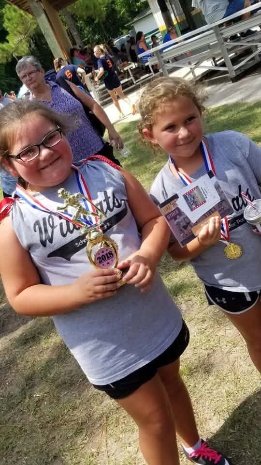 Two girls in grey Wildcats shirts wearing medals, one holding a small trophy