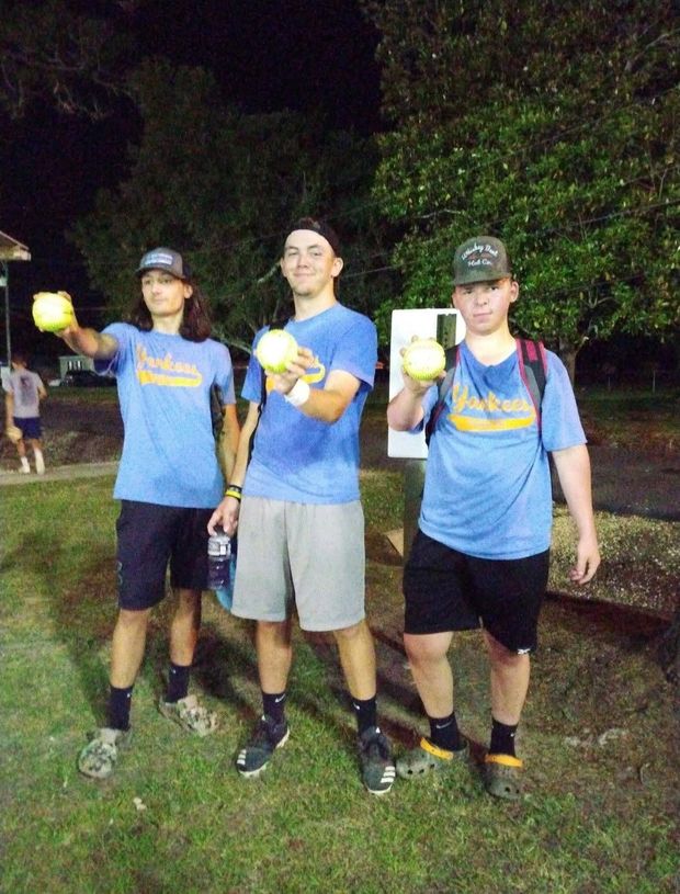Three teenagers in blue Yankees shirts each holding up a softball at night